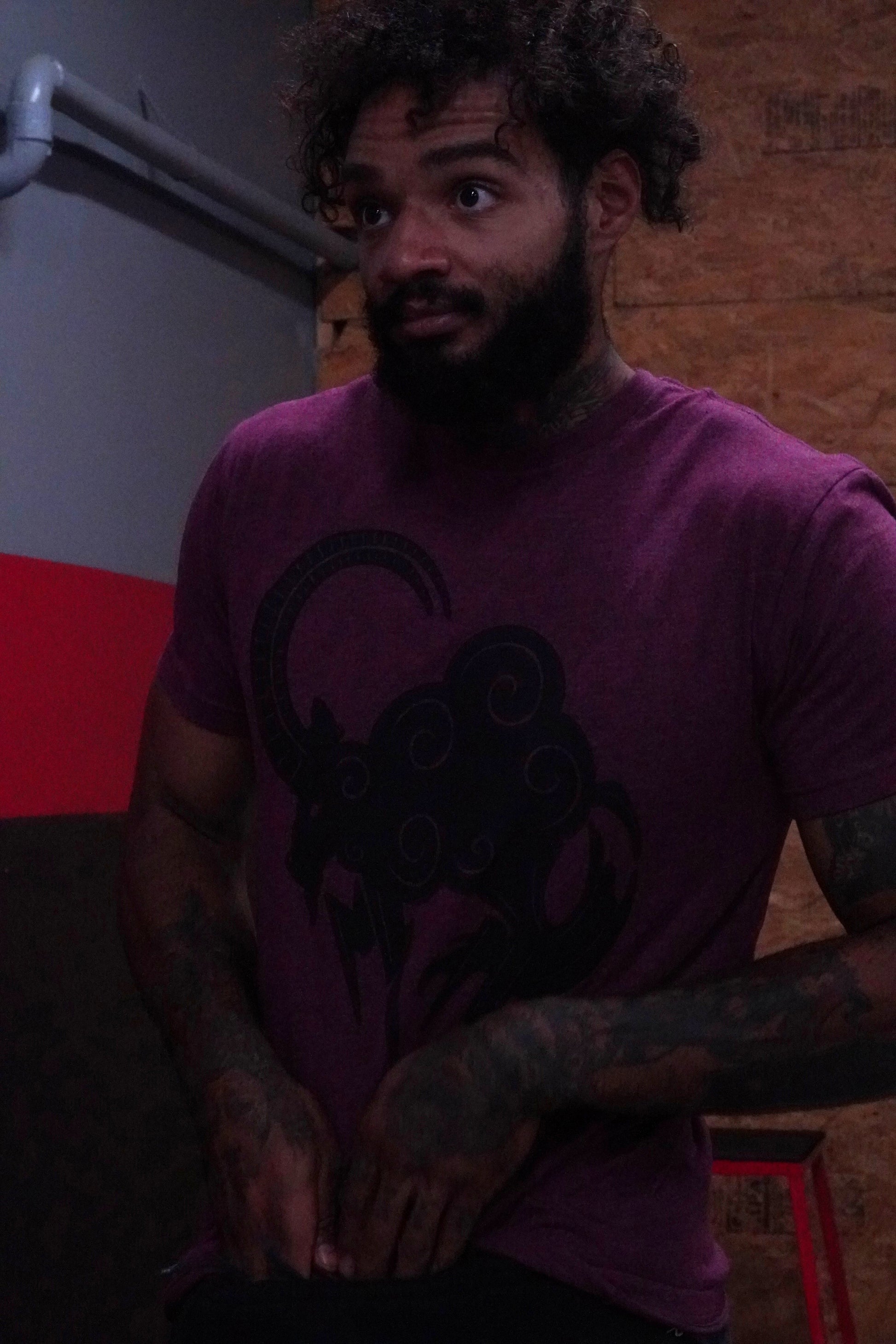 Man wearing a blood red t-shirt with a graphic design, standing against a red and gray wall.